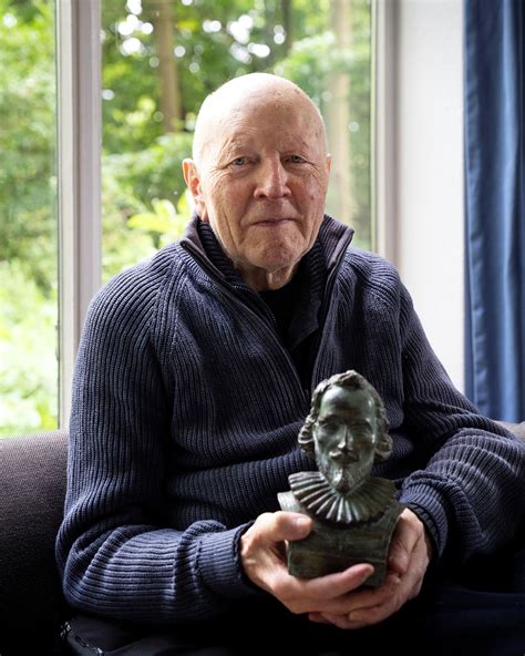 Een portret van Maarten 't Hart, staand in een weelderige tuin met boeken op de achtergrond.